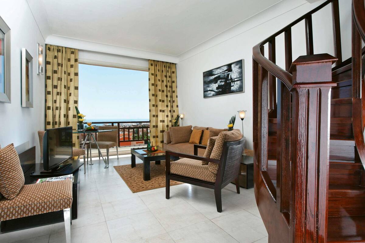Ein Wohnzimmer mit Sofa, Sessel, Fernseher und einem Essbereich mit Tisch und Stühlen. Eine Treppe ist rechts zu sehen.