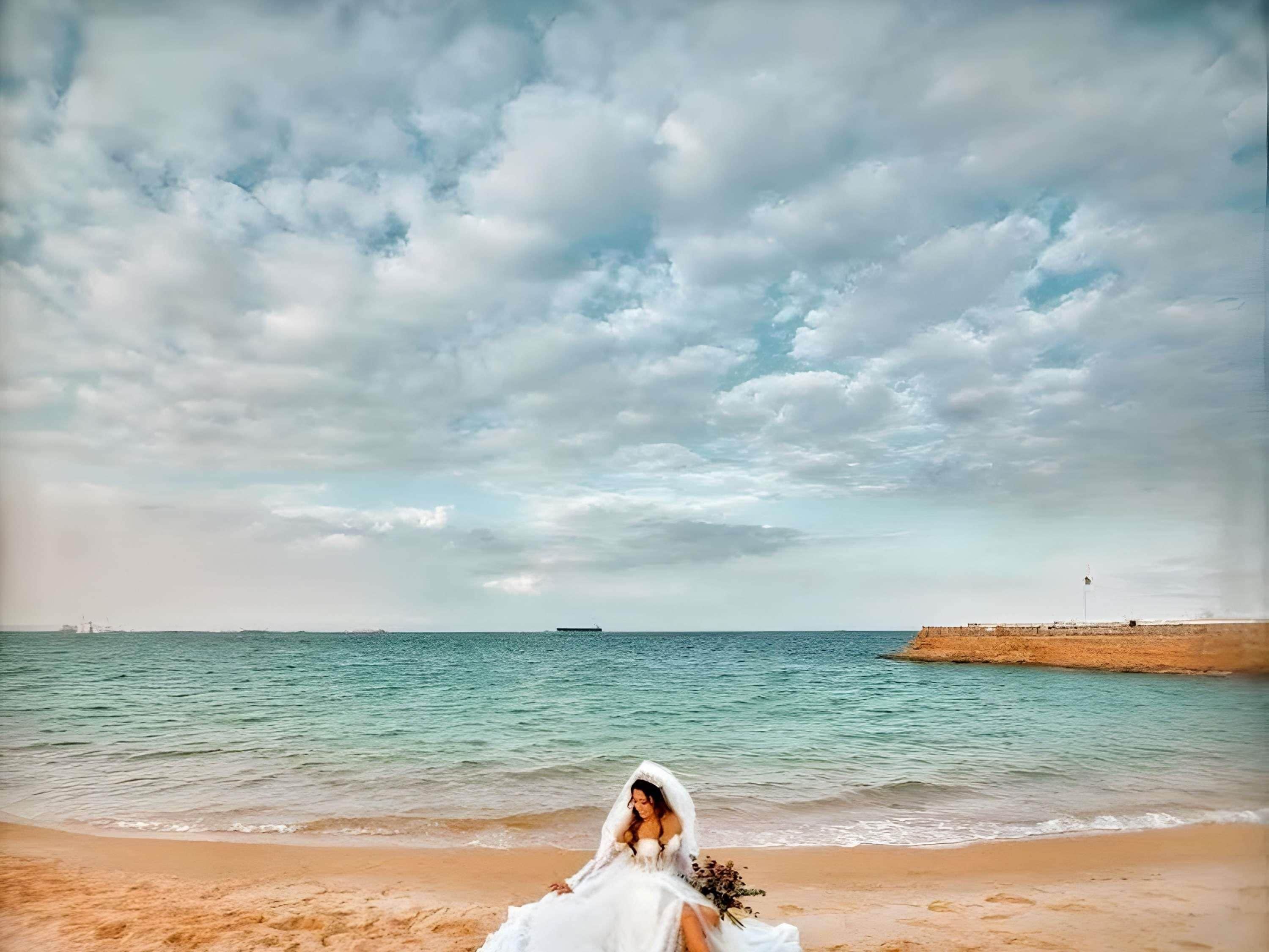Une scène de plage avec une personne vêtue d’une robe blanche, une plage de sable et une étendue d’eau.