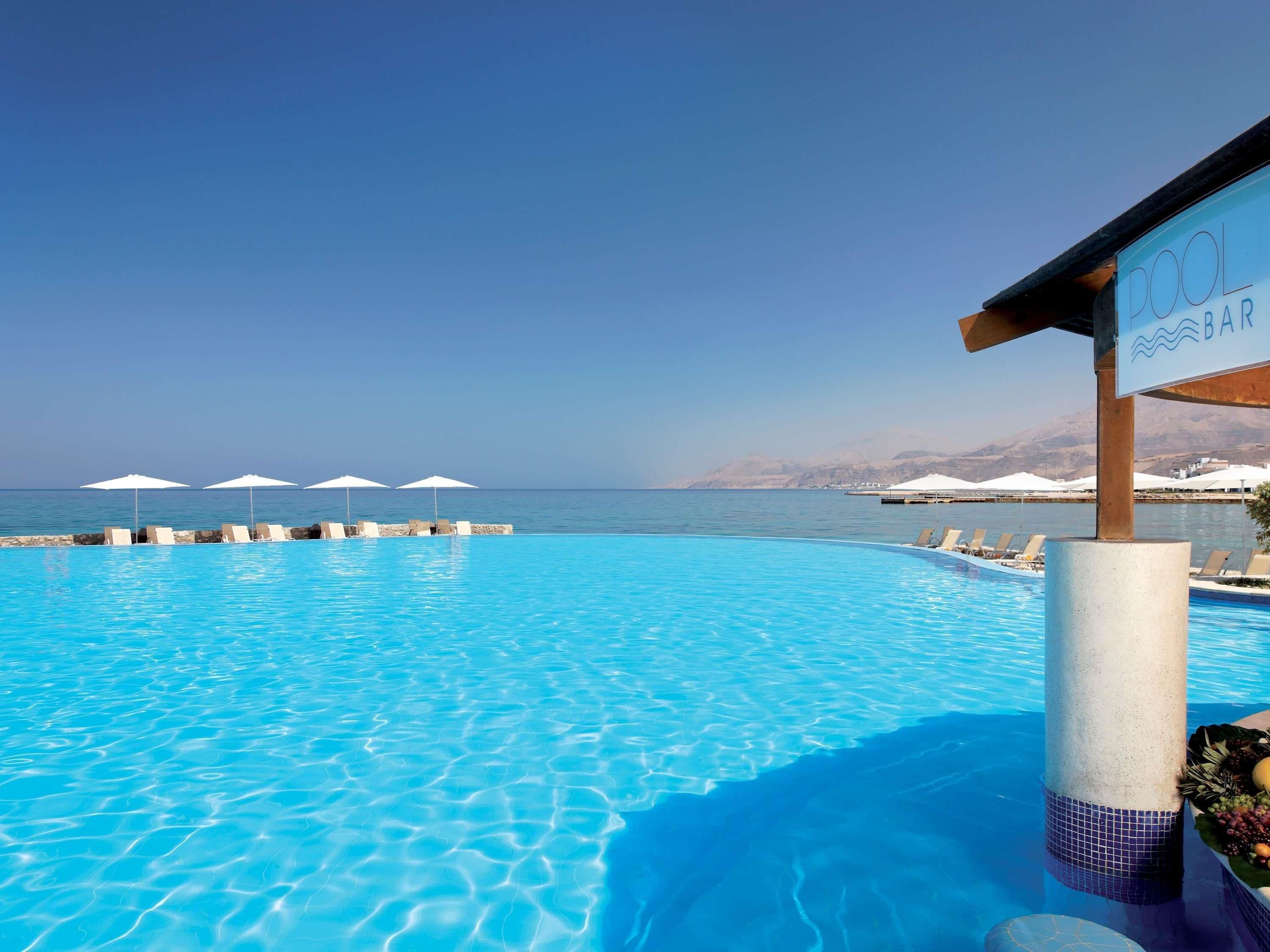 A clear blue pool with lounge chairs and umbrellas, a bar sign, and a view of the sea and mountains.