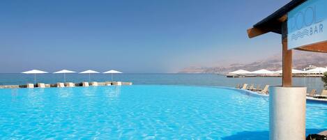 A clear blue pool with lounge chairs and umbrellas, a bar sign, and a view of the sea and mountains.