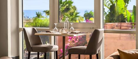 A dining area with a table set for two, two chairs, a view of the ocean, and tropical plants.