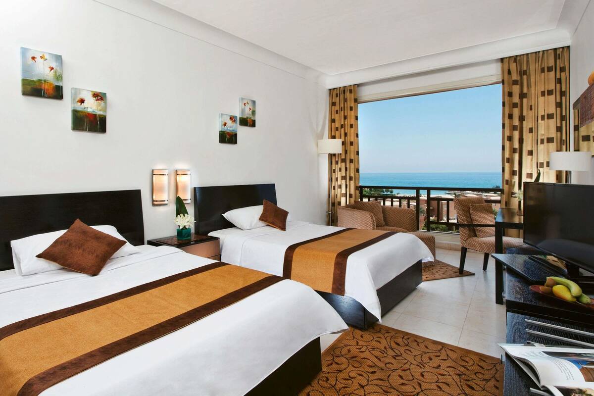 Ein Hotelzimmer mit zwei Betten, einem Fernseher, einem Esstisch mit Obst und einem Balkon mit Meerblick.