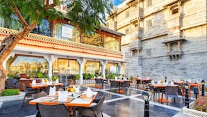 An outdoor dining area with tables and chairs, a tree, and a building with ornate architecture in the background. - Shiv Niwas Palace by HRH Group of Hotels (Udaipur)