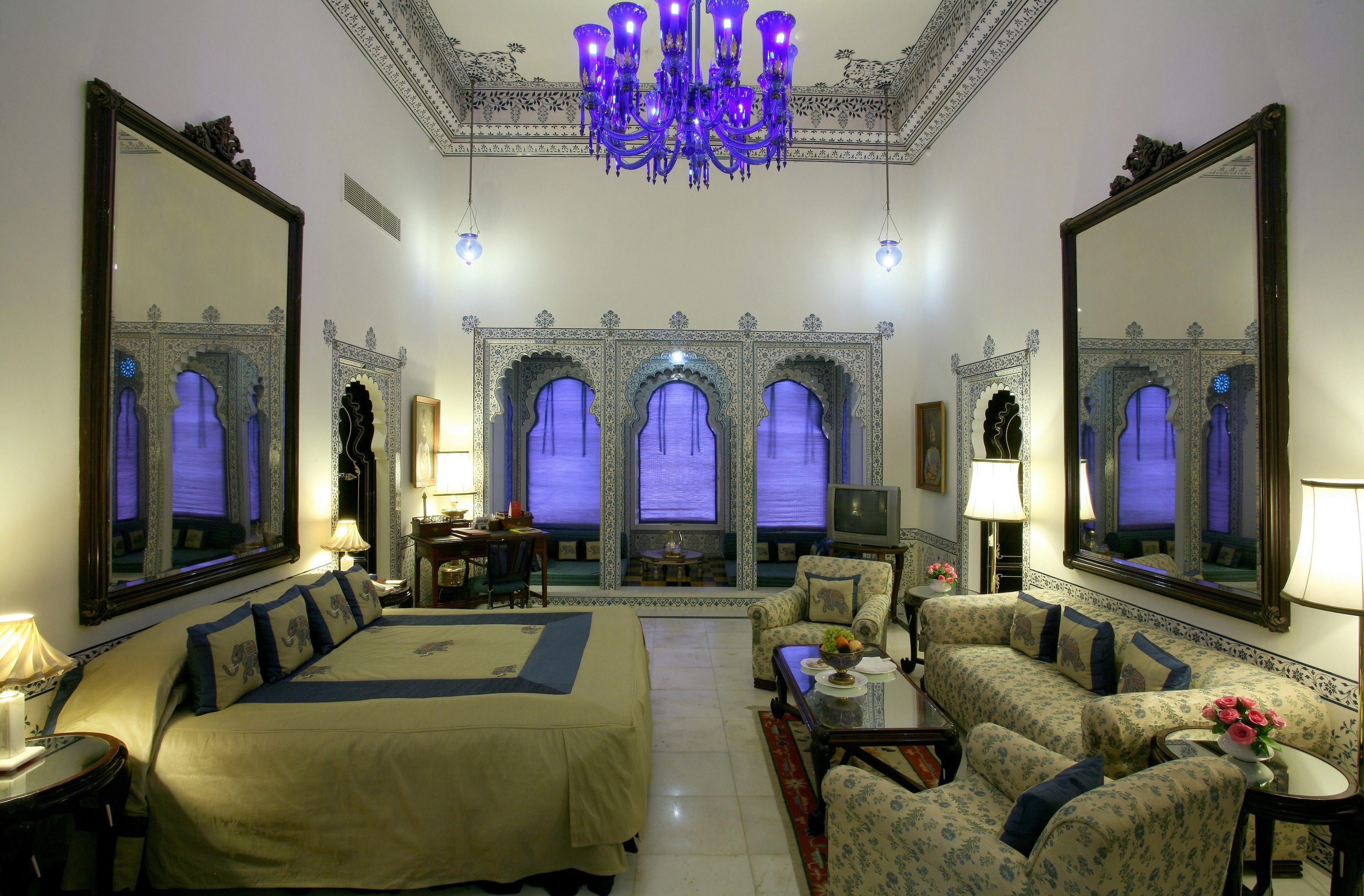 Quarto de hotel espaçoso com uma cama grande, vários espaços de estar e decoração requintada, incluindo um candelabro e paredes com espelhos.