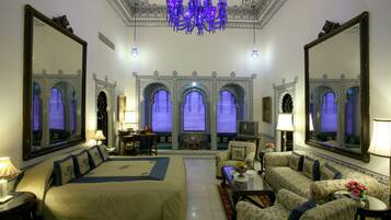 Quarto de hotel espaçoso com uma cama grande, vários espaços de estar e decoração requintada, incluindo um candelabro e paredes com espelhos.