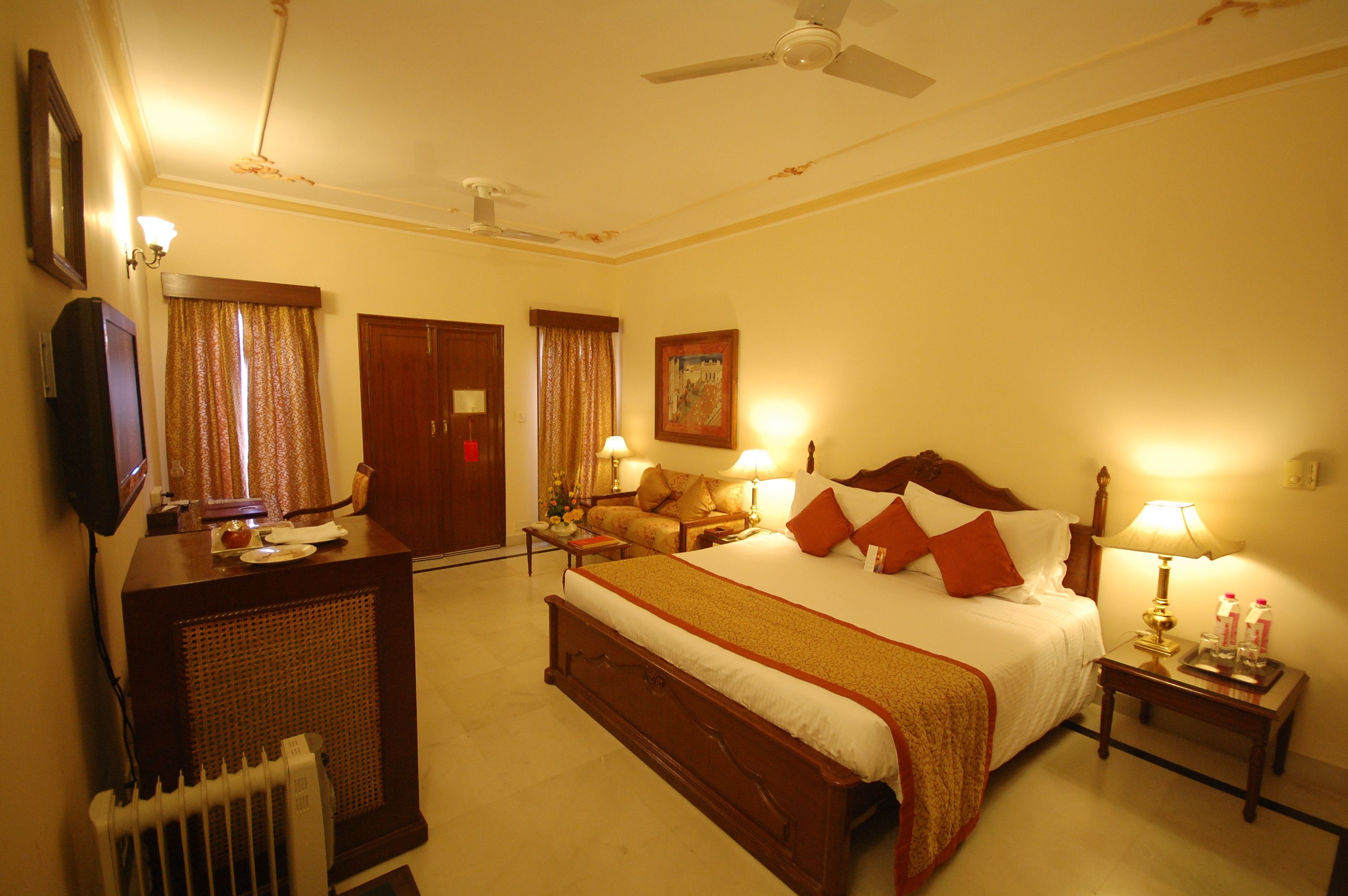 Une chambre d’hôtel avec un grand lit, un bureau, une chaise, un radiateur, un ventilateur de plafond et une table de chevet avec une lampe et une bouteille.