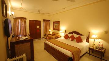 Une chambre d’hôtel avec un grand lit, un bureau, une chaise, un radiateur, un ventilateur de plafond et une table de chevet avec une lampe et une bouteille.