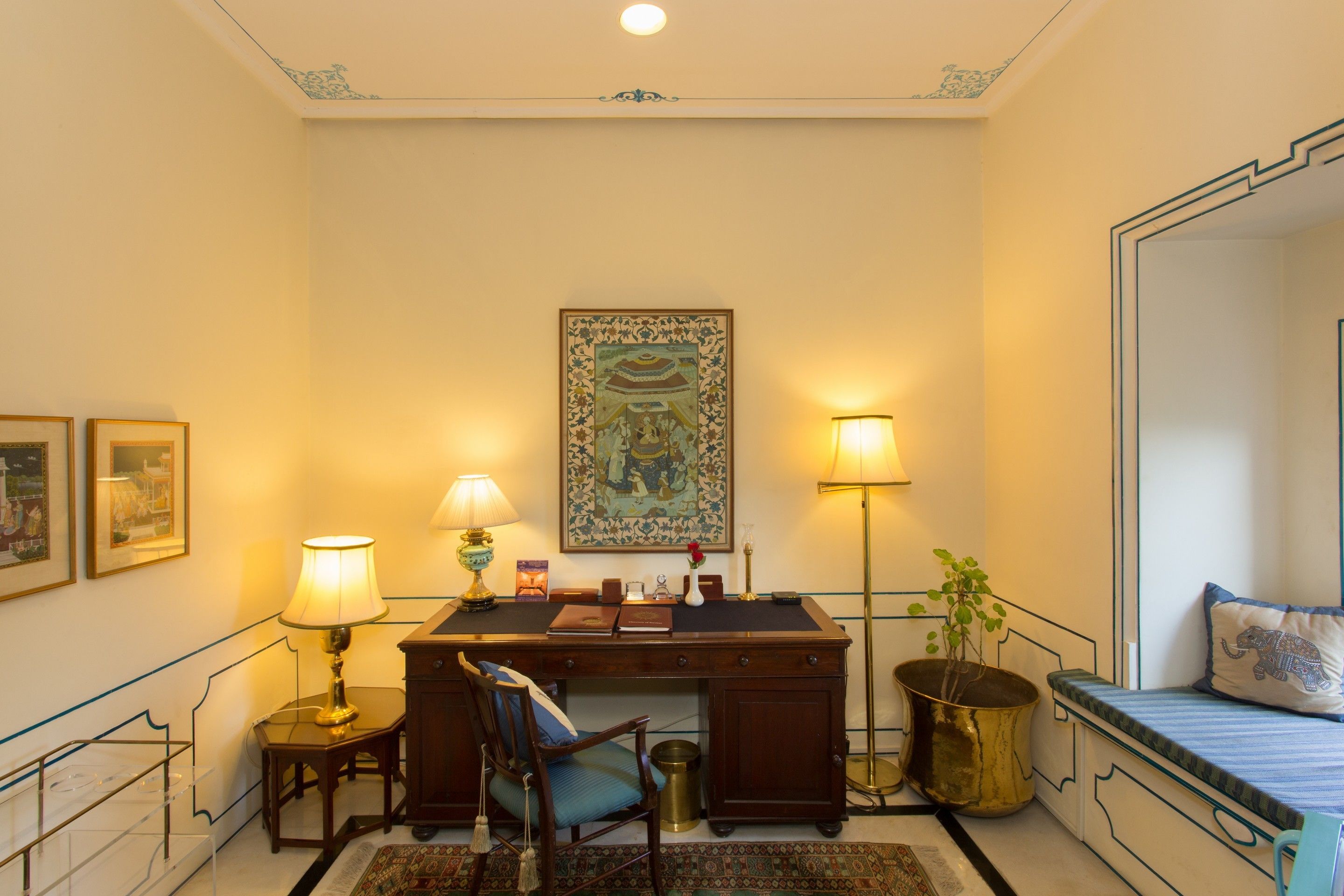A room with a desk, chair, lamp, framed artwork, and a bed with a pillow.