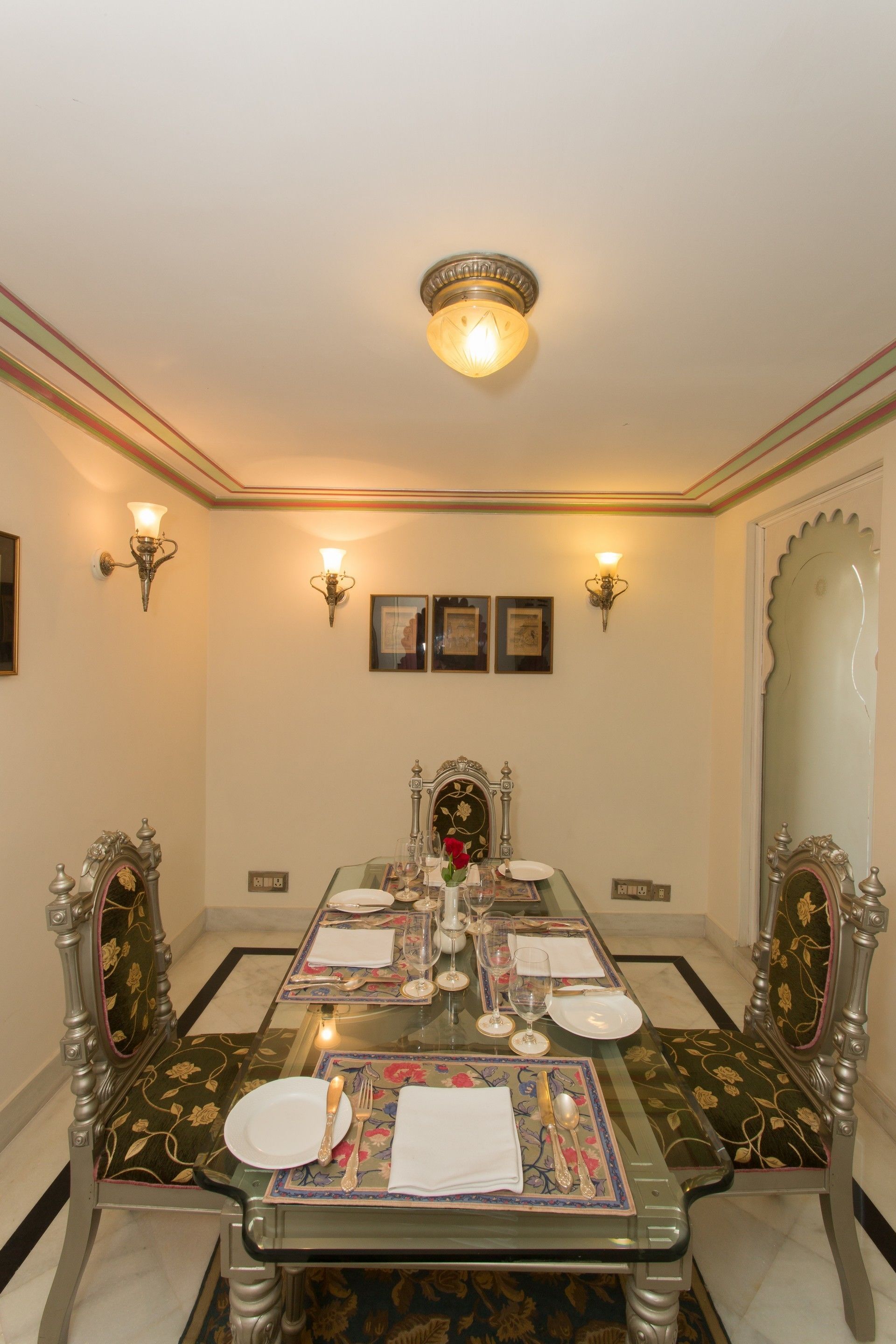 Comedor con una mesa de vidrio dispuesta para cuatro, sillas ornamentadas, apliques de pared y cuadros enmarcados.