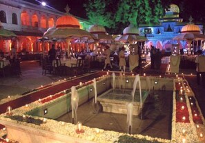 An outdoor dining area with tables and umbrellas, a decorative pool, and a building with arches in the background. - Shiv Niwas Palace by HRH Group of Hotels (Udaipur)