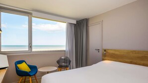 A hotel room with a bed, a chair, a small table, a lamp, and a view of the beach. - ibis Le Touquet Thalassa (Le Touquet-Paris-Plage)