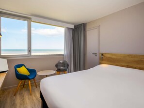 A hotel room with a bed, a chair, a small table, a lamp, and a view of the beach. - ibis Le Touquet Thalassa (Le Touquet-Paris-Plage)