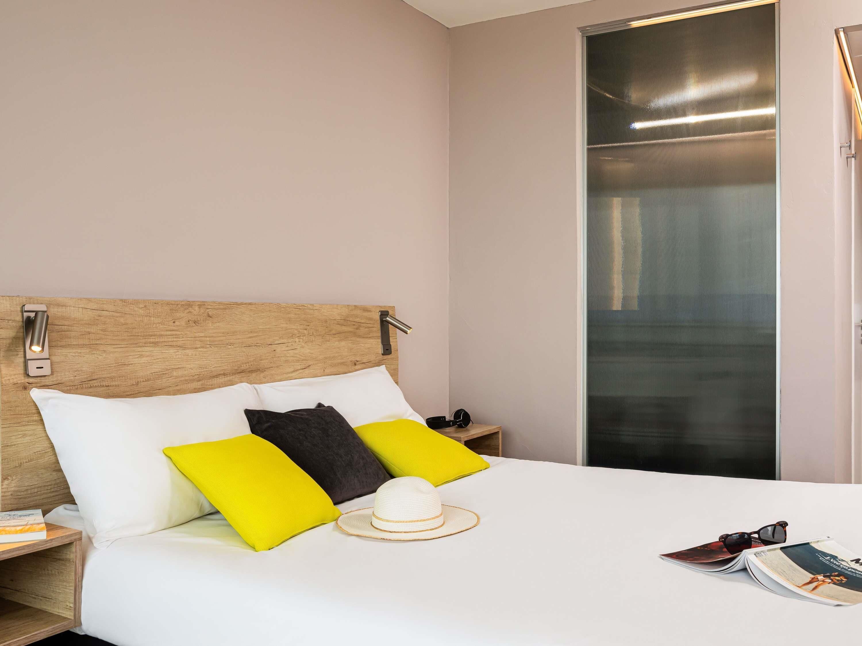 A neatly made bed with white and yellow pillows, a wooden headboard, and a nightstand with a book and a hat.