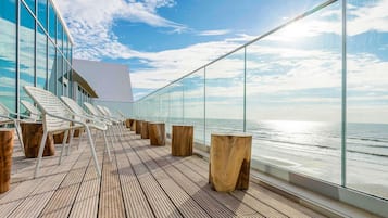 Une terrasse avec un plancher en bois, des chaises blanches et des tabourets en bois, offrant une vue sur l’océan.