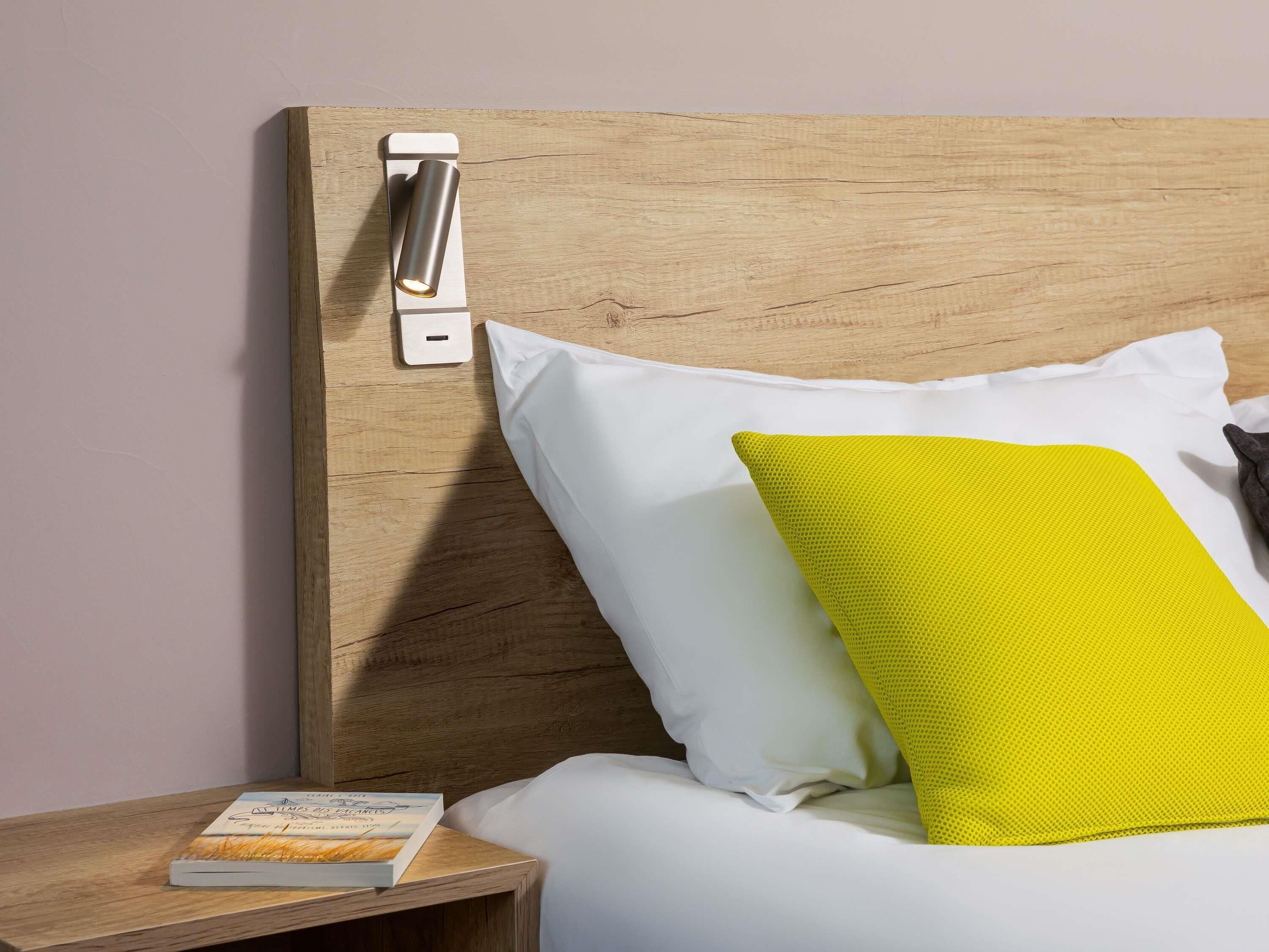 A neatly made bed with white and yellow pillows, a bedside table with a book, and a wall-mounted light switch.