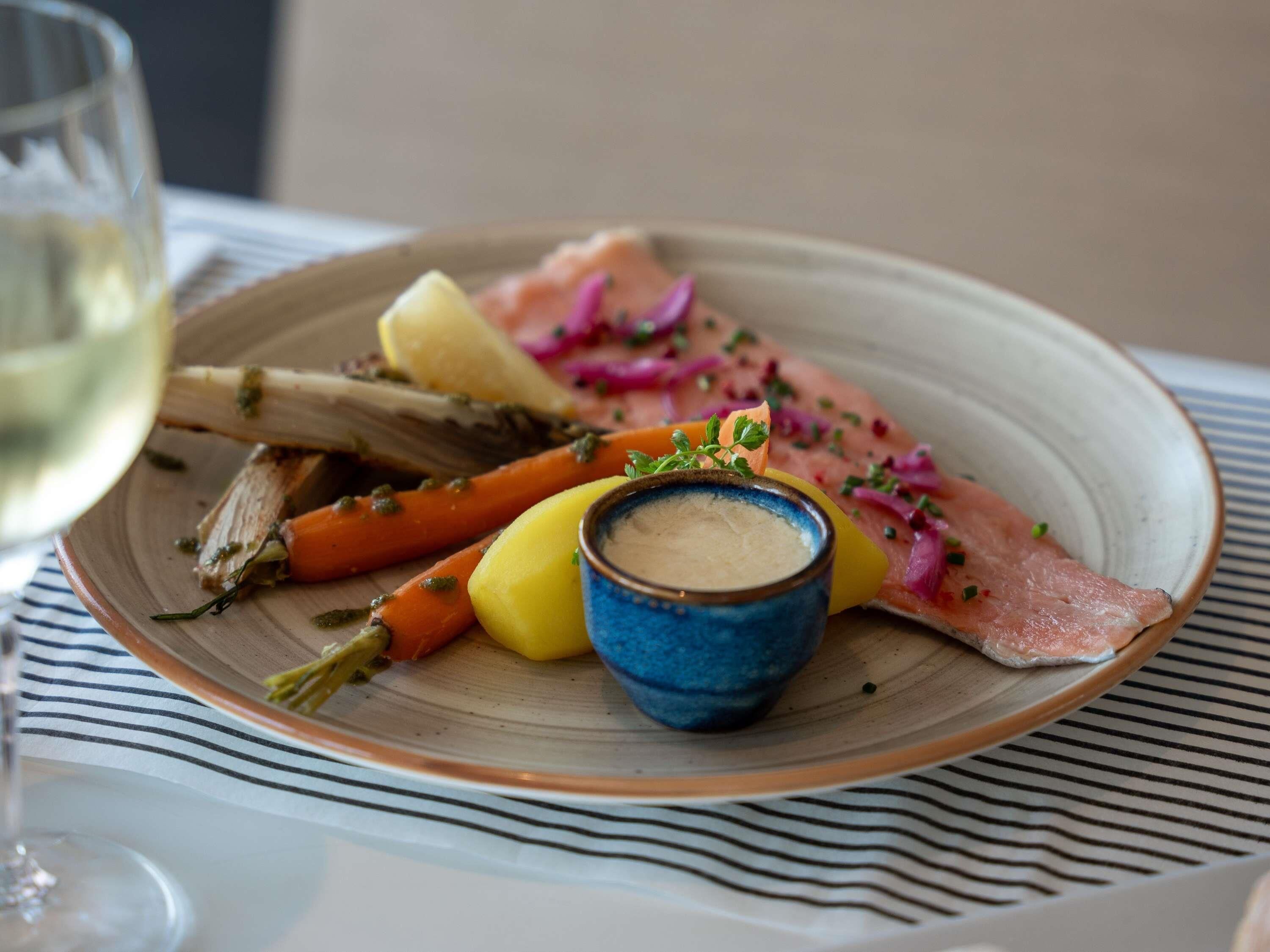 A plate with salmon, vegetables, and a small bowl of sauce on a striped tablecloth.