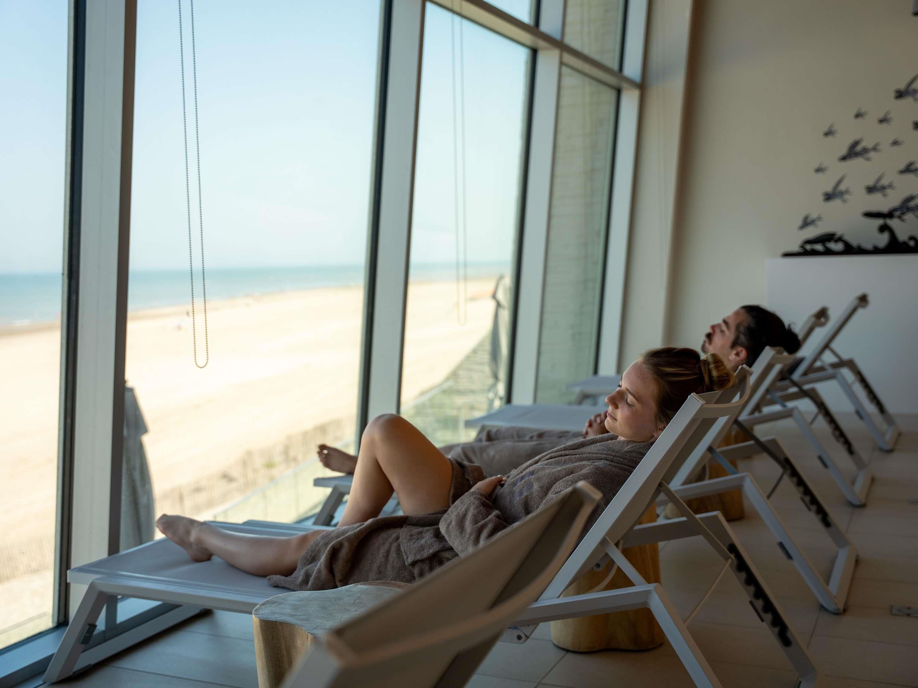 A room with large windows overlooking a beach, equipped with reclining chairs and a massage table.