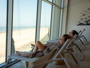 Habitación con grandes ventanales con vista a la playa, equipada con sillones reclinables y una camilla de masaje.