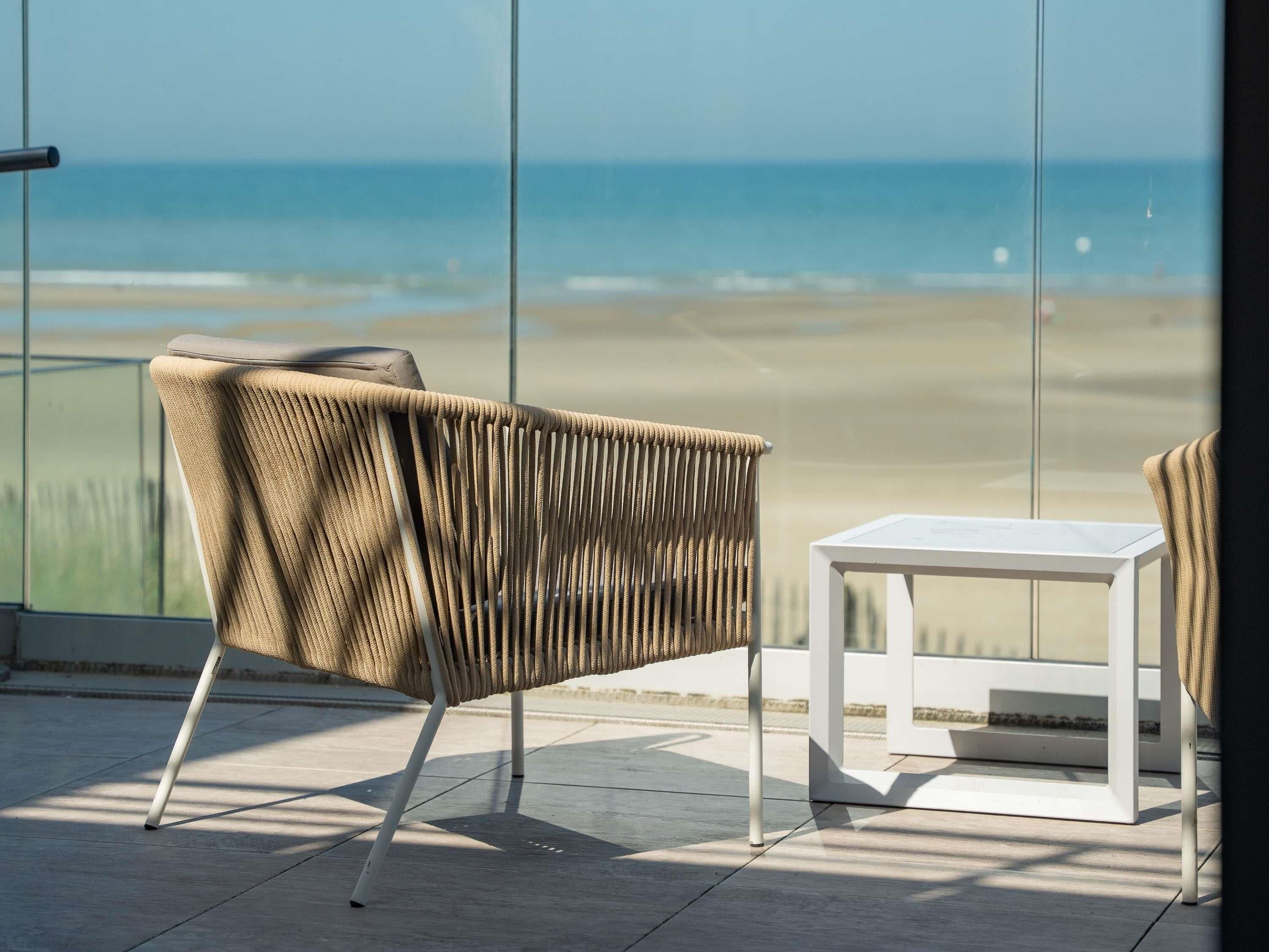 A wicker chair with a cushion and a small white table on a balcony with a view of the beach.