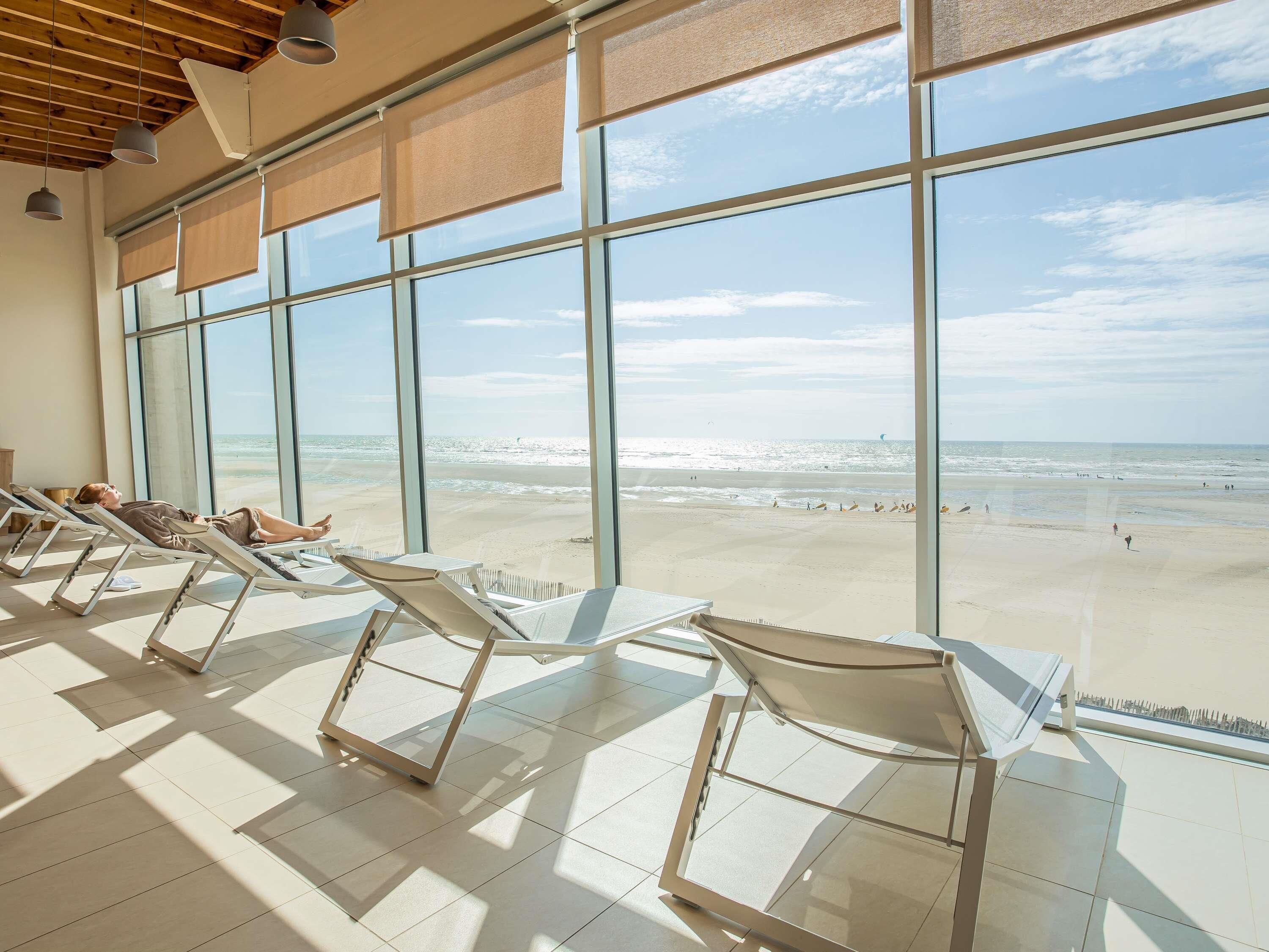 Une chambre située en bord de mer, dotée de grandes fenêtres, de chaises longues et offrant une vue sur l’océan.