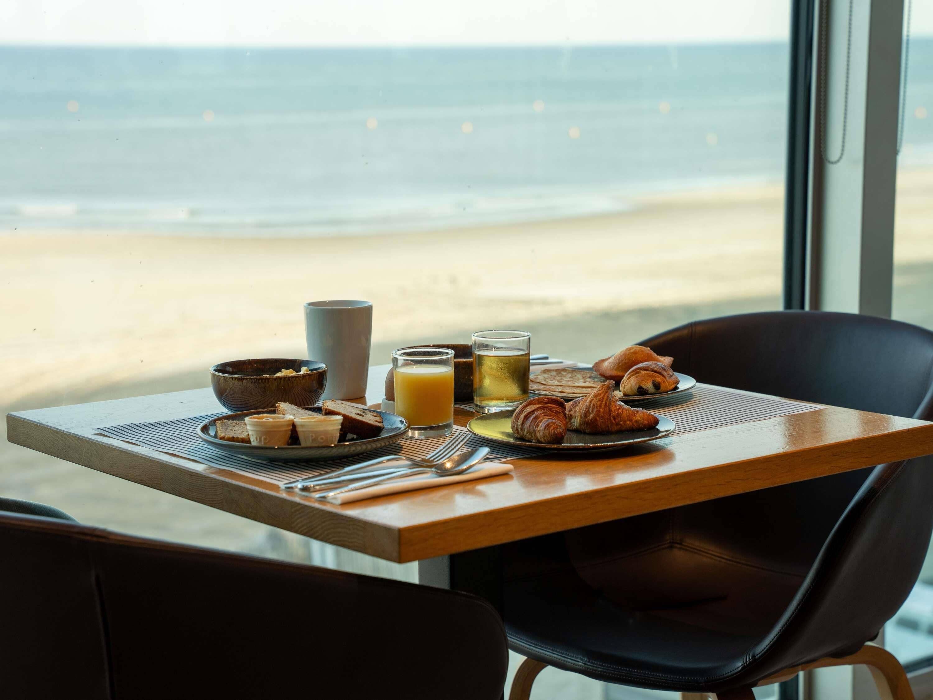 Una tavola per la colazione con croissant, una ciotola, un bicchiere di succo e una tazza, con vista sulla spiaggia.