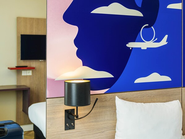 Habitación de hotel con una lámpara de pared, una cama con almohada blanca, una maleta y un televisor en la pared.