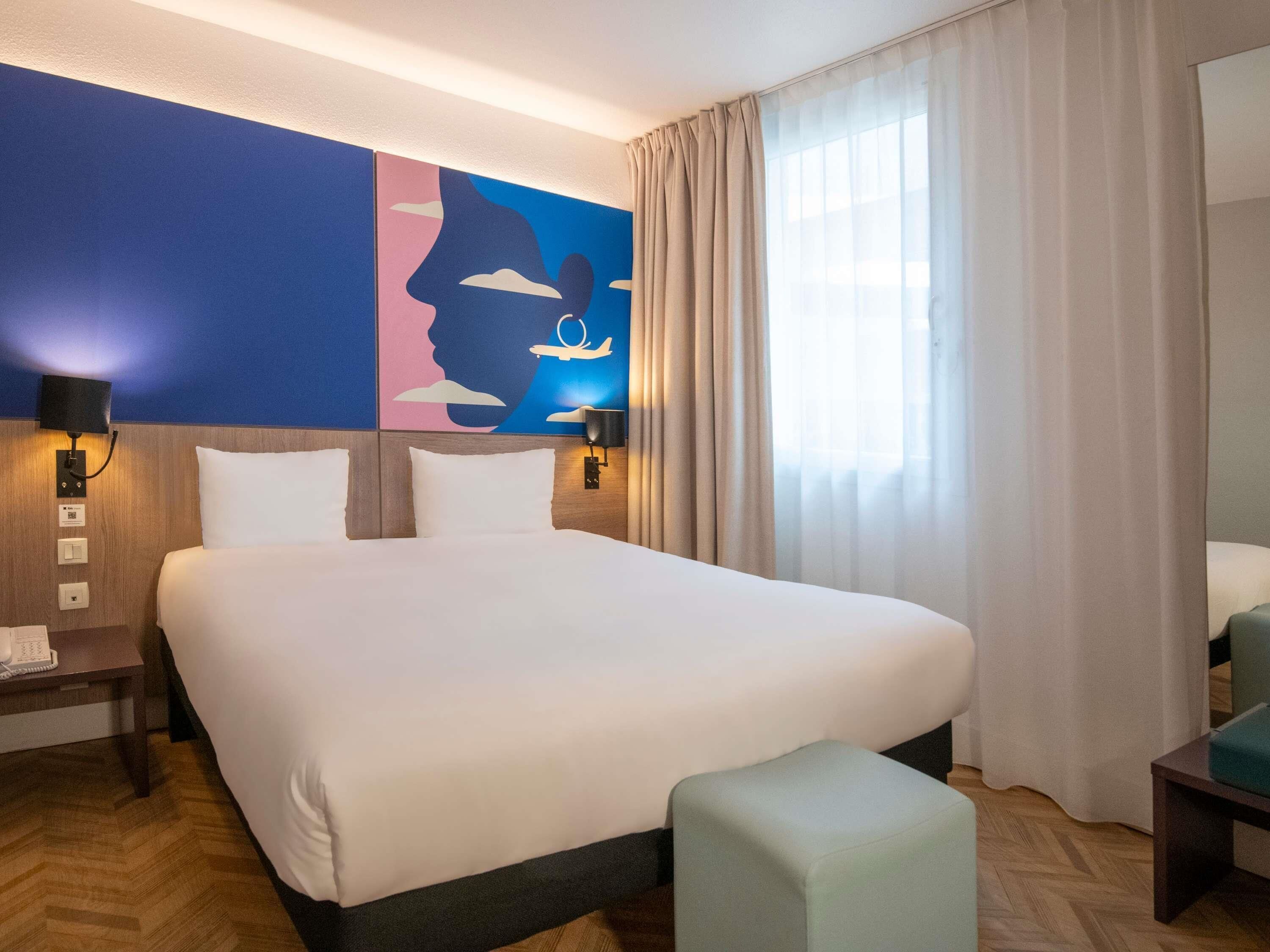 A hotel room with a large bed, two bedside tables, a desk, a chair, and a painting on the wall.