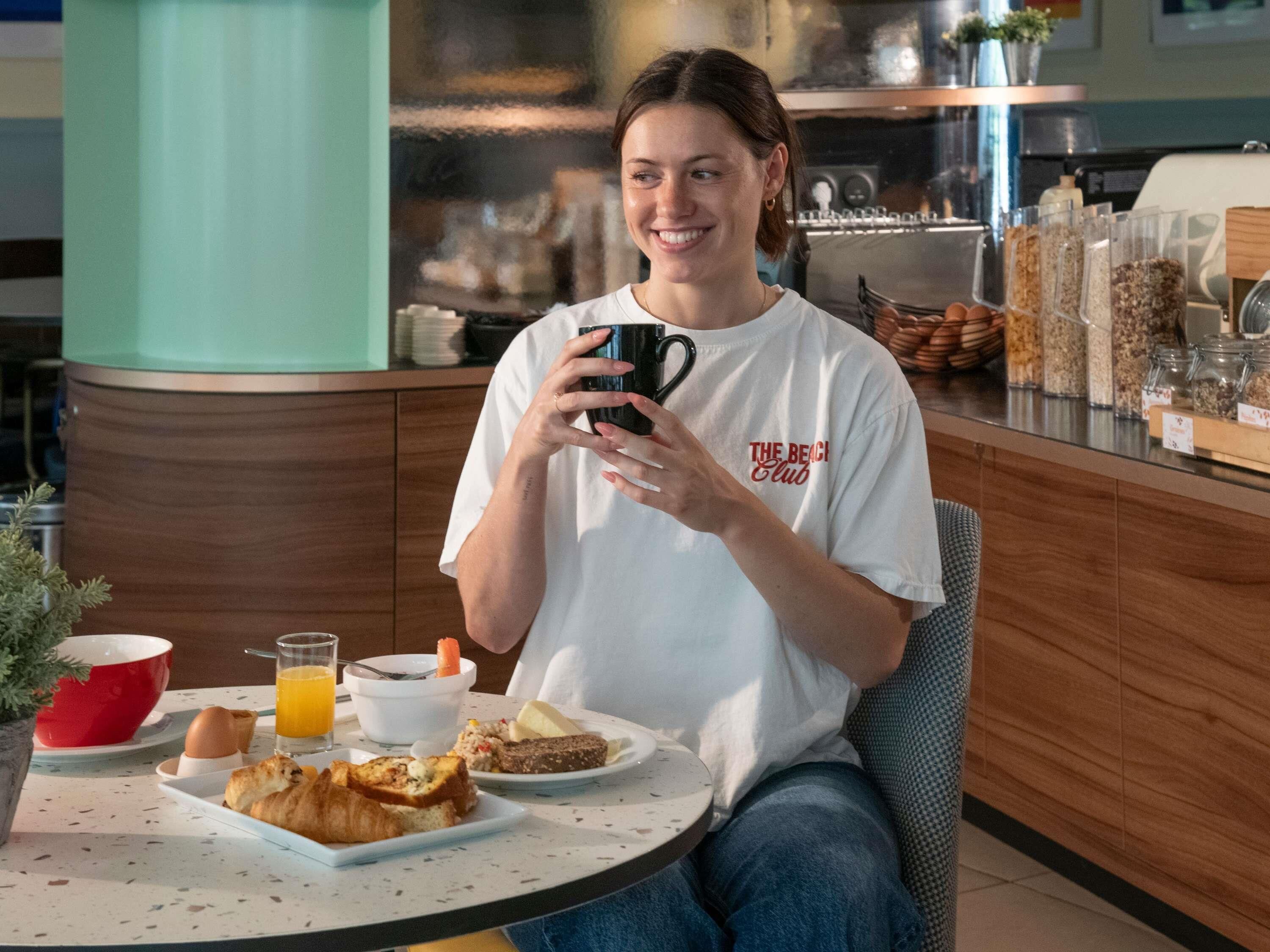 Una persona sentada a una mesa con comida para el desayuno, una taza de café y una cocina al fondo.