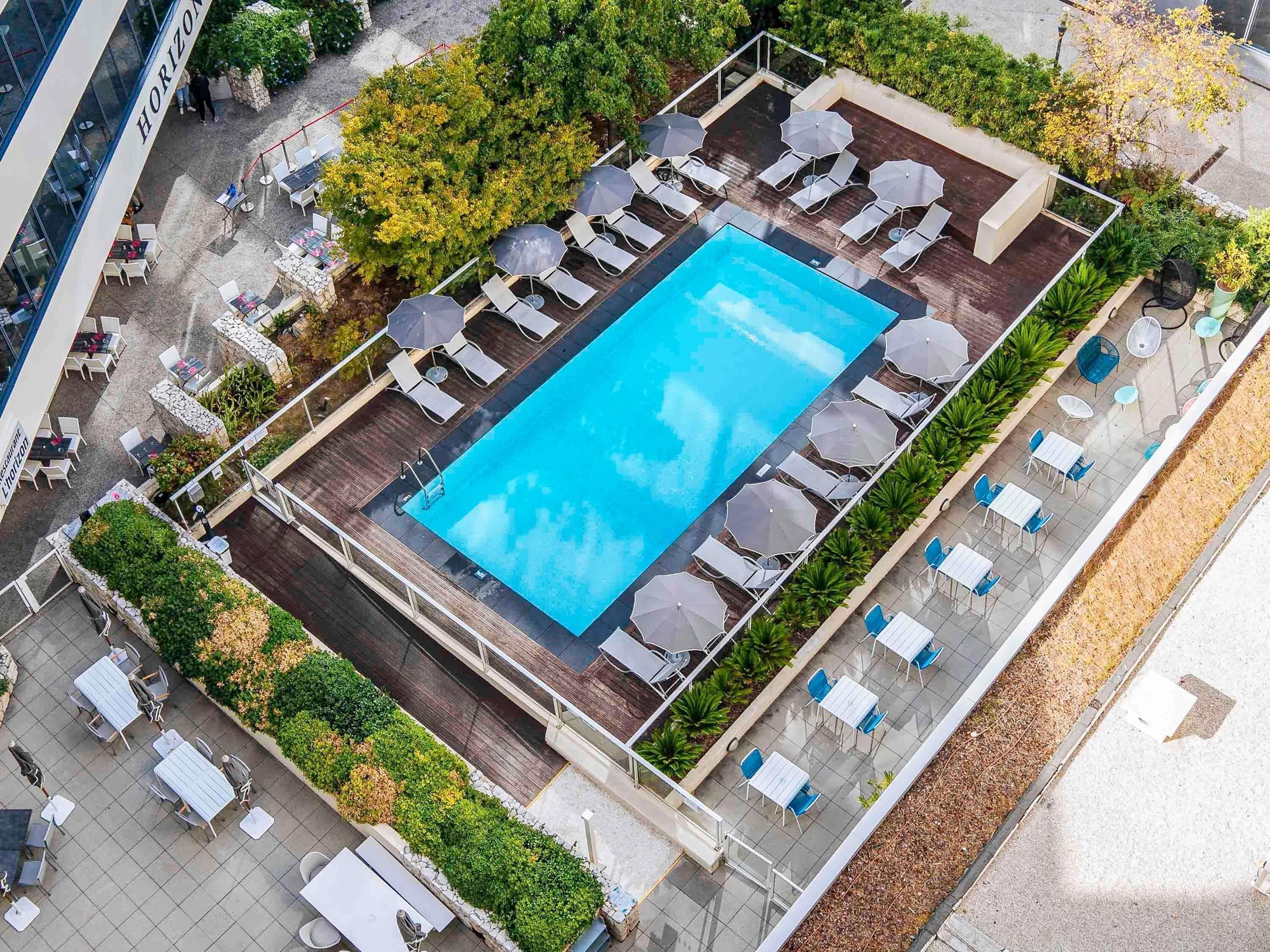 Vista aérea de una zona de piscina en la azotea con sillones de descanso y sombrillas, rodeada de vegetación y un área de comedor.