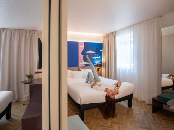 A hotel room with a bed, a bedside table, a chair, a small plant, and a wall with a graphic design. - ibis Styles Nice Aéroport Arenas (Nice)