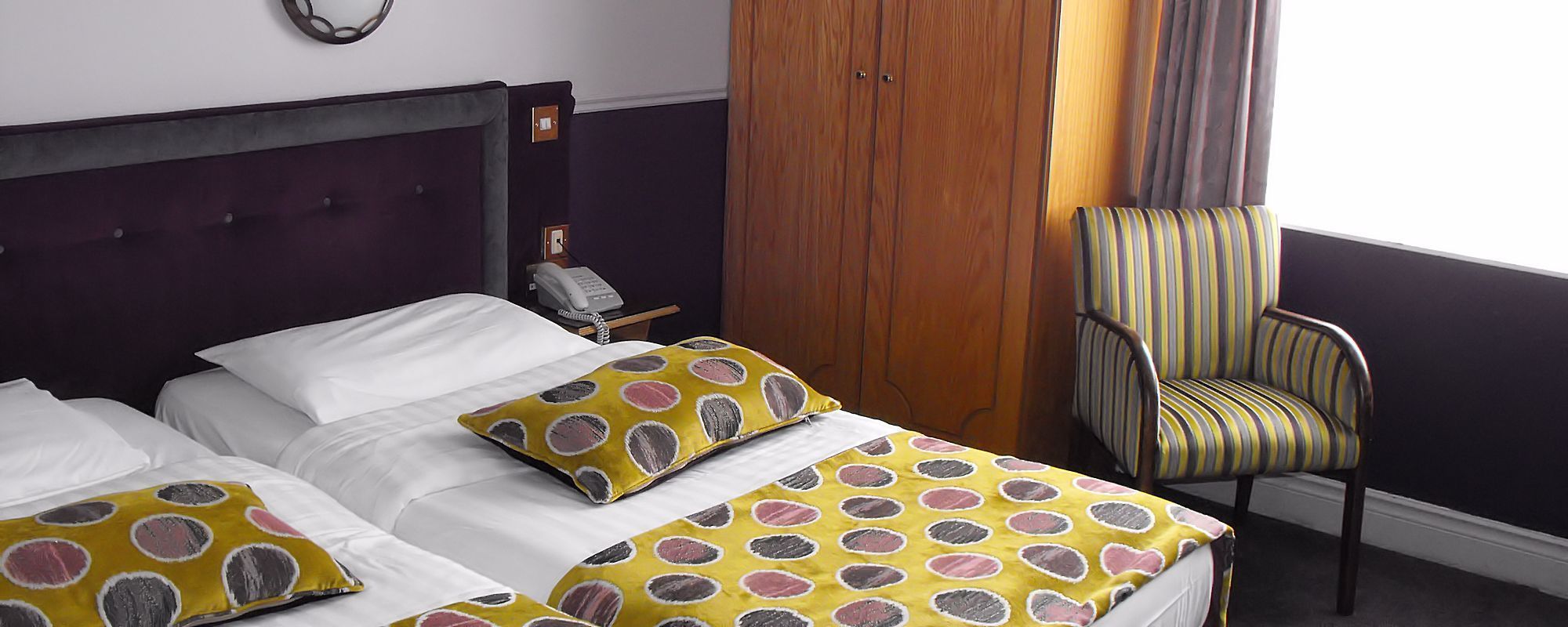Une chambre d’hôtel avec deux lits, une armoire en bois, une chaise à rayures et un téléphone sur la table de chevet.