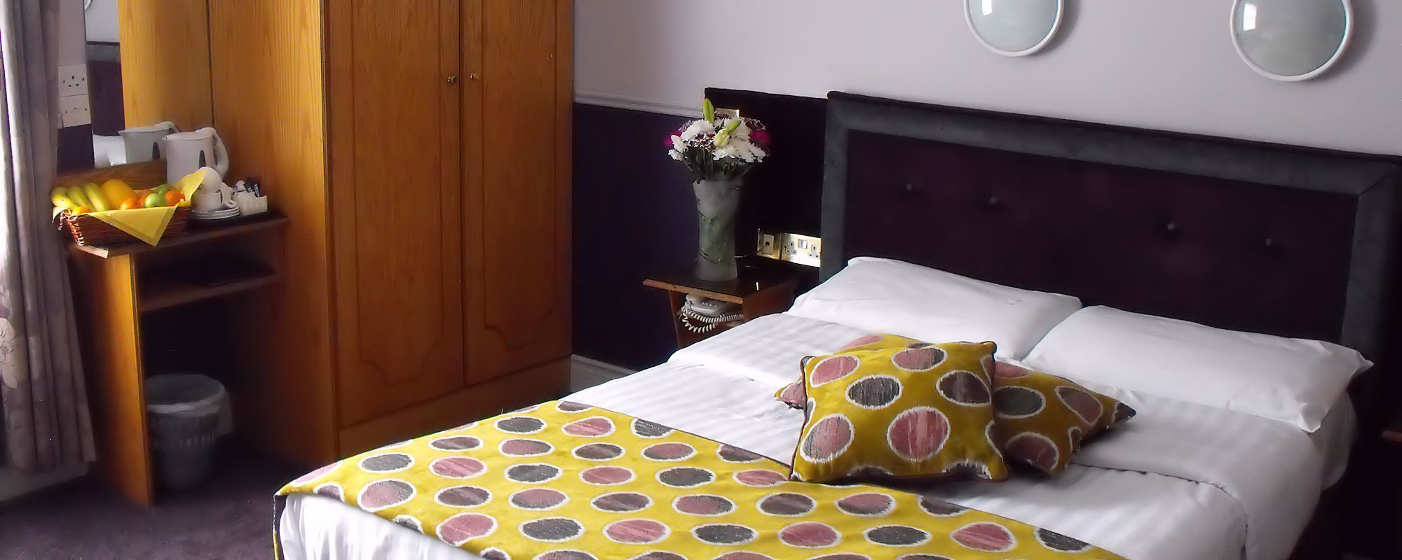 Una cama bien hecha con un edredón de lunares, un armario de madera y un florero con flores en una mesita de noche.
