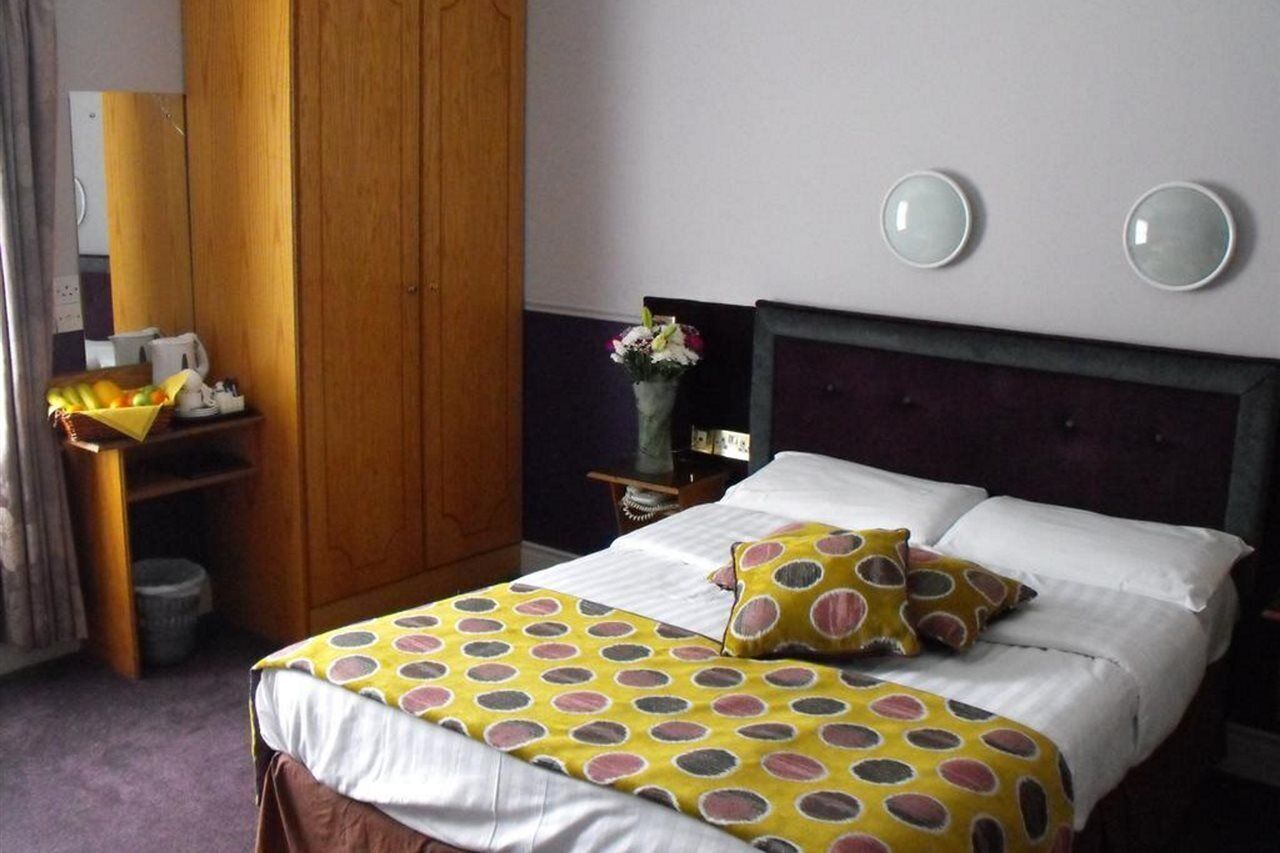 A hotel room with a bed, a wooden wardrobe, a nightstand with a vase of flowers, and a mirror on the wall.