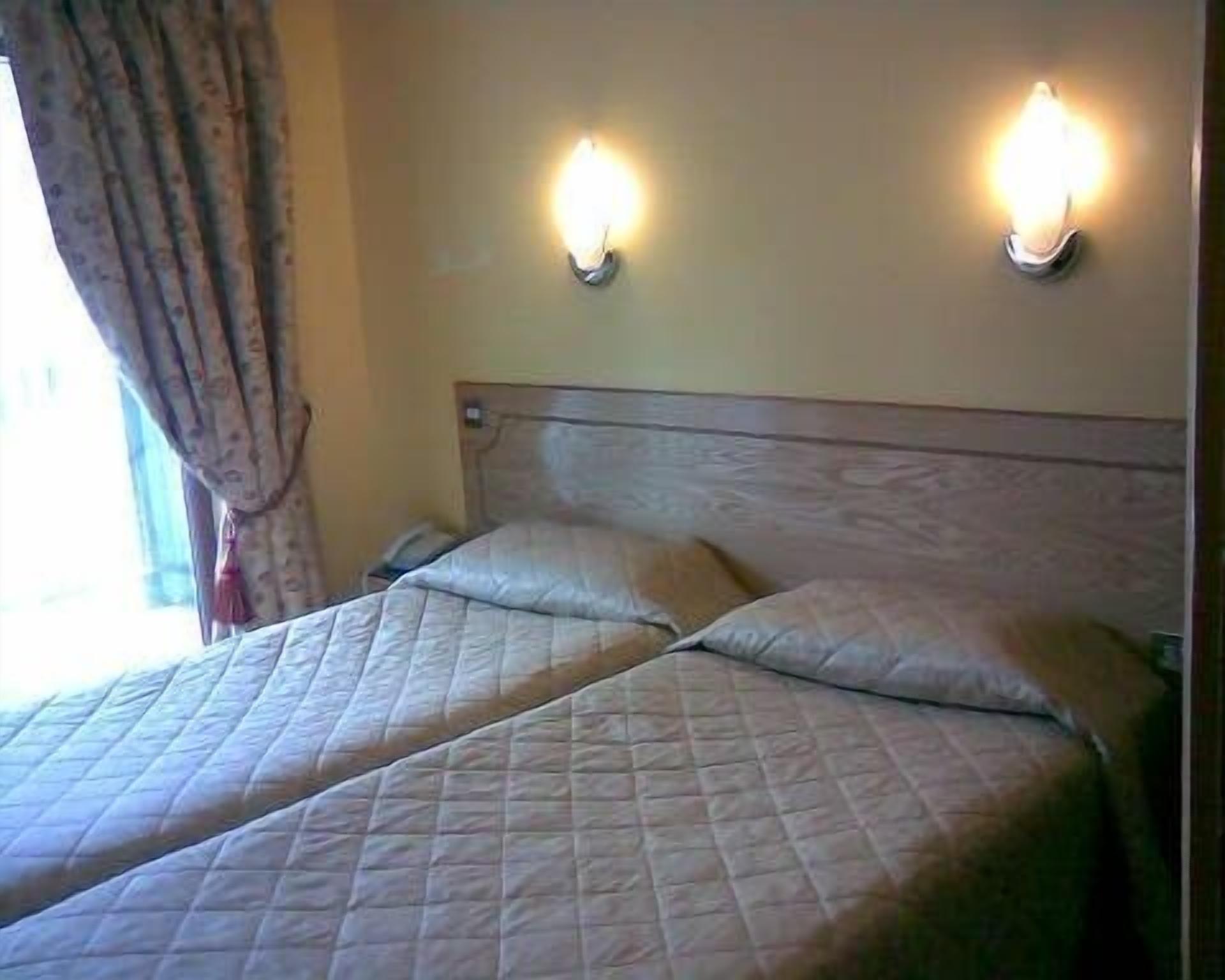 A hotel room with two beds, each with a headboard and a bedside table. There are wall-mounted lights above the beds.