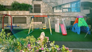 Un parque infantil con columpios, tobogán y estructuras para escalar, frente a un edificio de piedra con balcón.