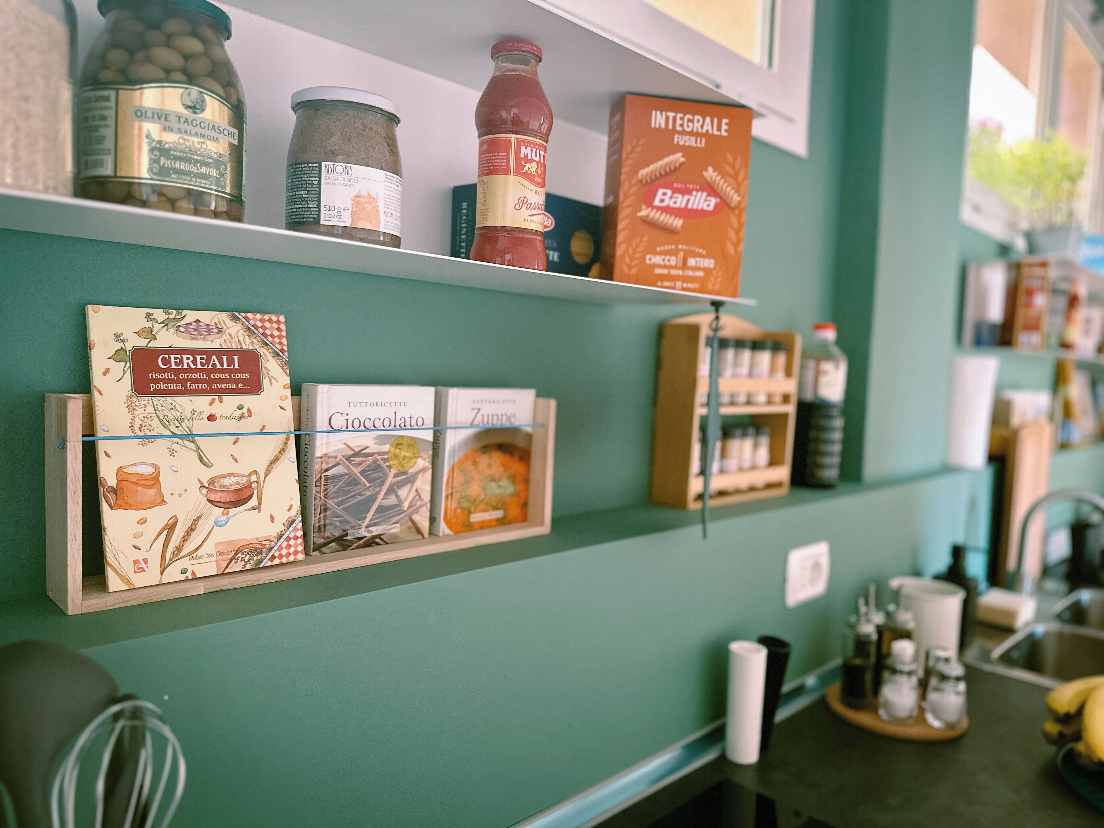 A kitchen shelf with jars of olives, a bottle of ketchup, a box of Barilla pasta, and cereal boxes.