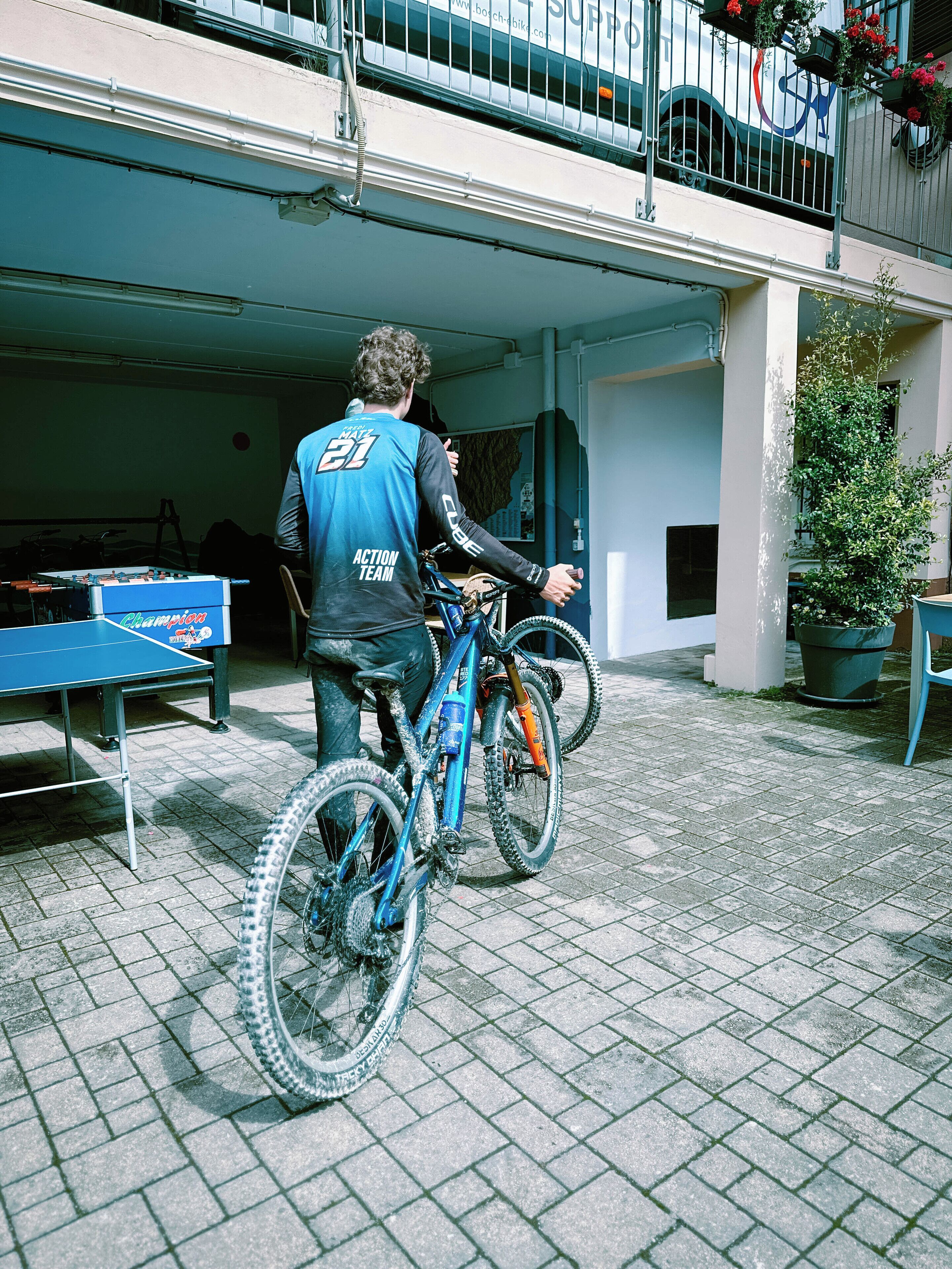 Una persona con una bicicletta di fronte a un edificio con un basculante, un tavolo da ping pong e una pianta in vaso.