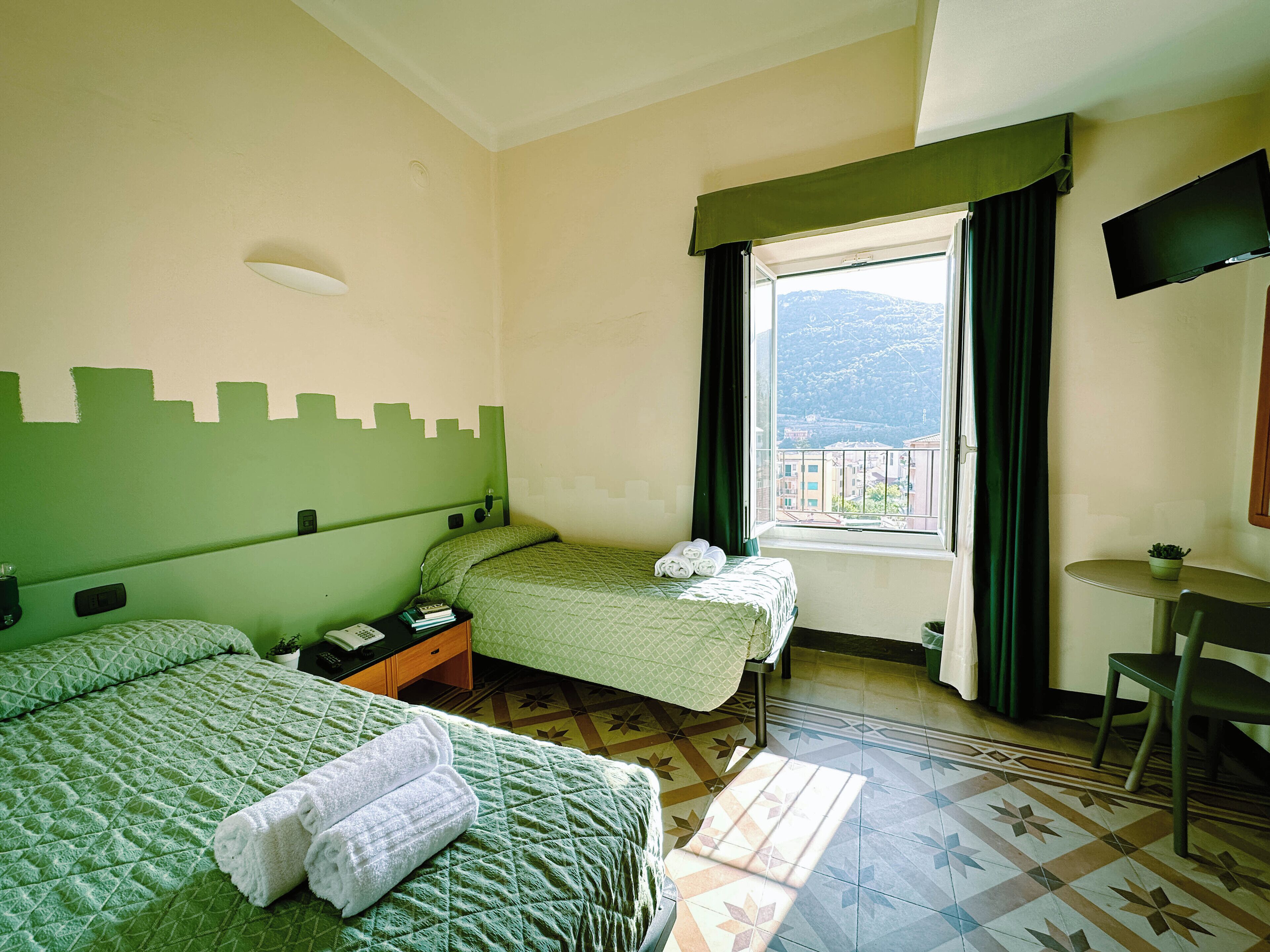 A hotel room with two beds, a desk, a chair, a TV, and a window with a view of buildings and mountains.