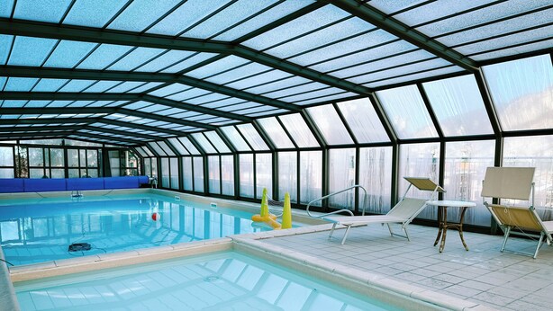 Una piscina interior con paredes de vidrio transparente, piso de baldosas y algunas sillas de descanso.