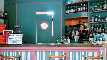 Angolo bar caratterizzato da una palette di colori verde acqua e rosa, con tre sgabelli, un bancone con una cassa e scaffali pieni di bottiglie.
