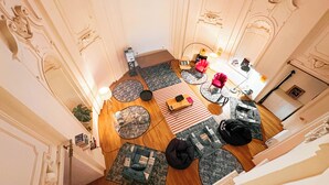 Amplia sala de estar con techo alto, ornamentación en las paredes y diversas opciones de asientos, incluyendo un sillón rojo, un sofá negro y varios tapetes redondos.
