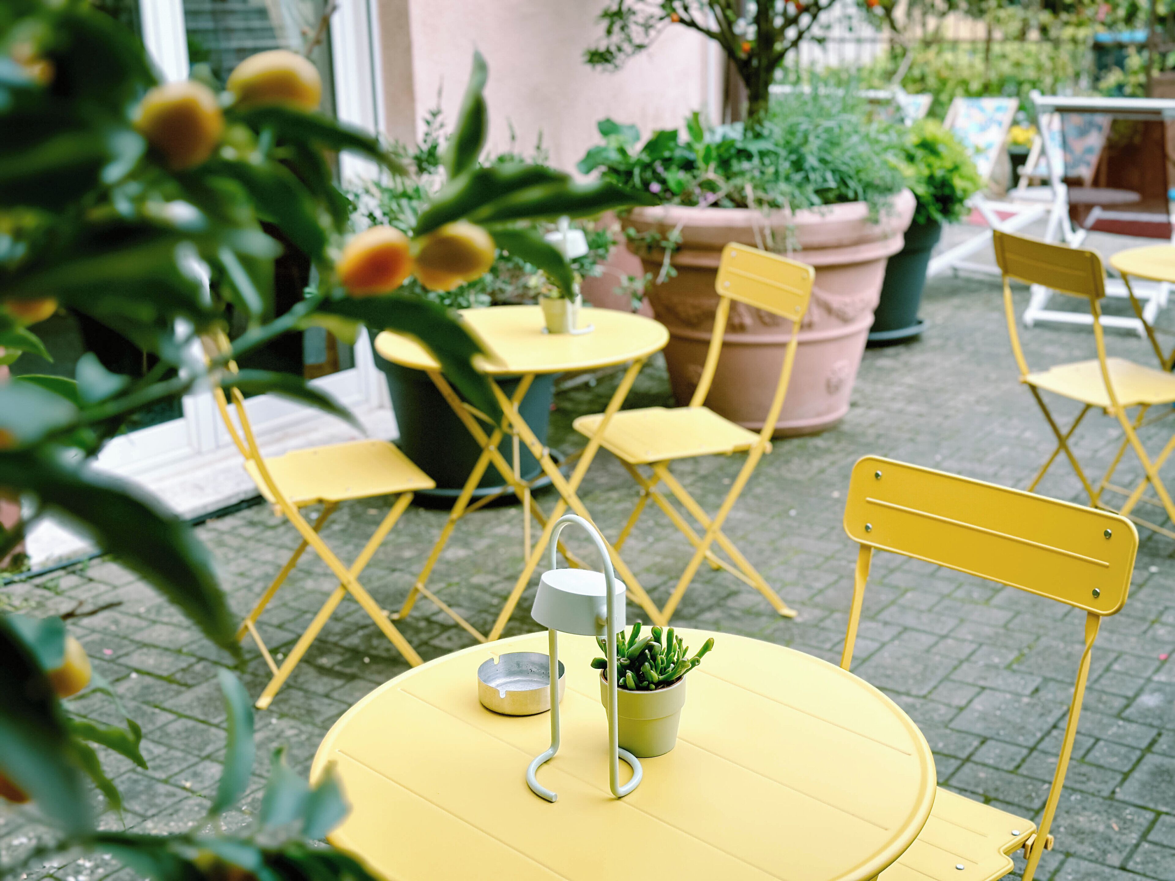 A yellow outdoor seating area with foldable chairs and a round table, surrounded by potted plants and a pink wall.