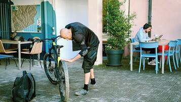 Una persona trabajando en una bicicleta al aire libre, con una mesa, sillas, una planta en maceta y un edificio al fondo.