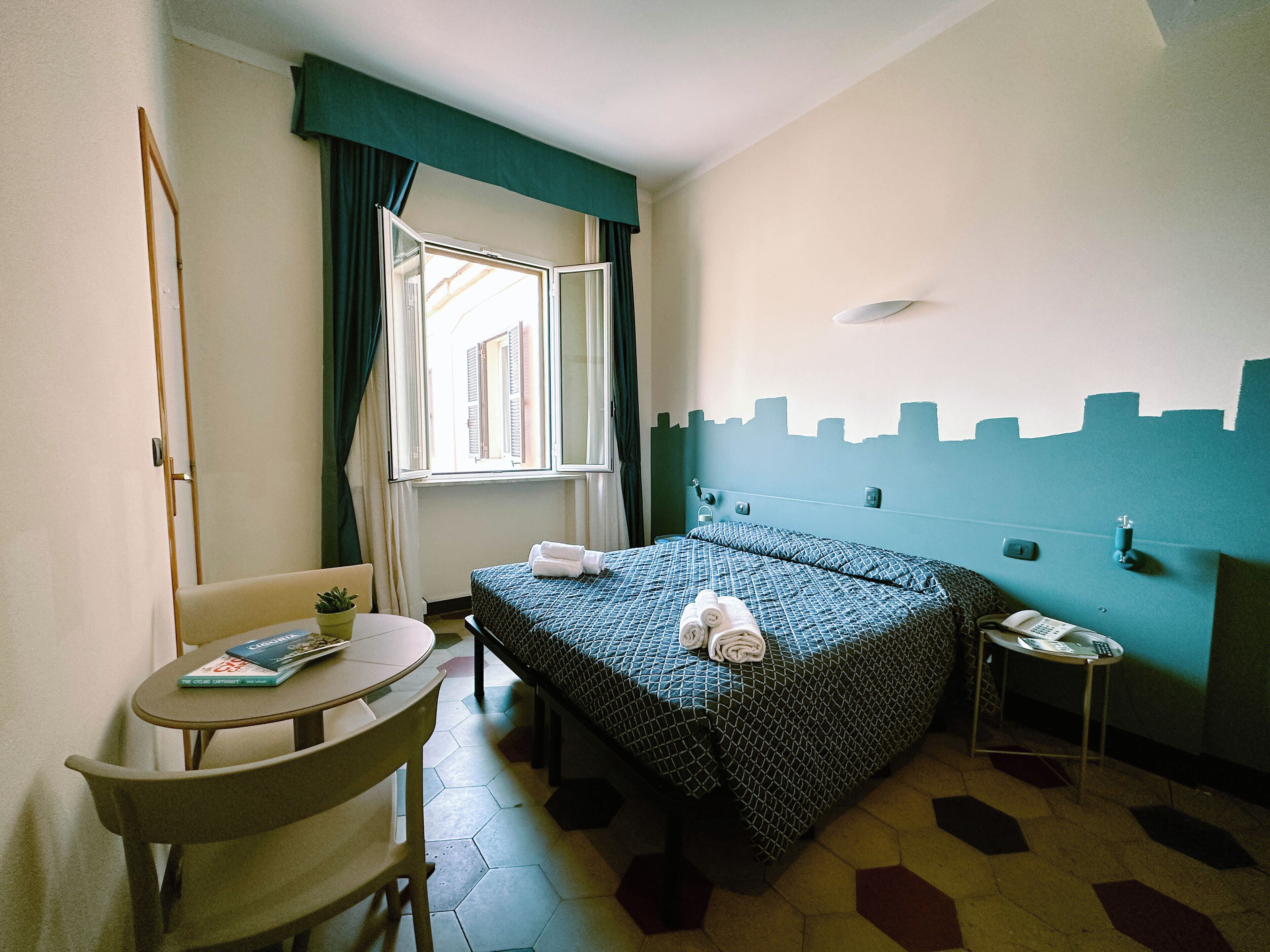 Une chambre d’hôtel avec un lit, une petite table et une chaise, une fenêtre avec des rideaux et un mur orné d’une fresque représentant un paysage urbain.