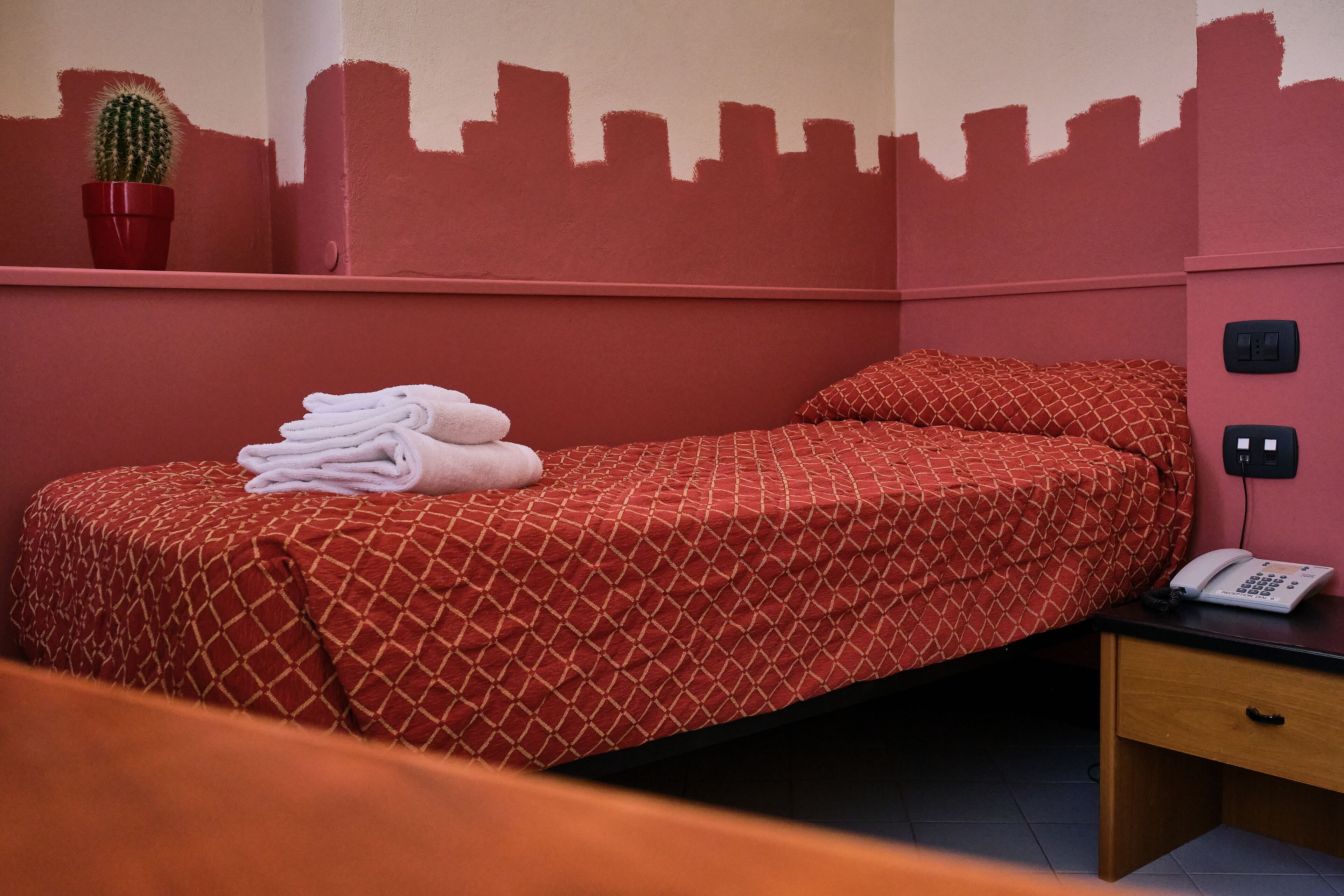 A single bed with a patterned red bedspread, a pair of folded white towels, a cactus in a red pot, and a cityscape silhouette on the wall.
