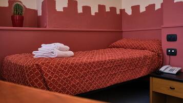 A single bed with a patterned red bedspread, a pair of folded white towels, a cactus in a red pot, and a cityscape silhouette on the wall.