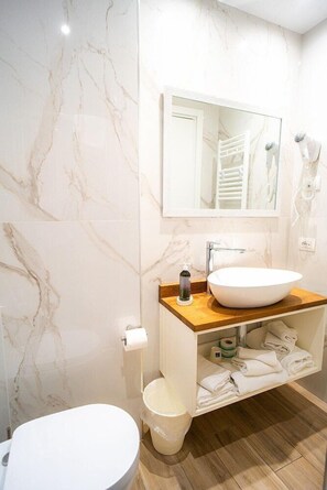 A bathroom with a white marble wall, a wooden vanity, a white sink, a toilet, and a mirror. - Continental (Pietra Ligure)