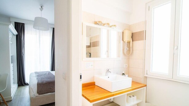 Une chambre d’hôtel avec un lit, une table de chevet en bois, un lavabo blanc, un miroir et une fenêtre avec des rideaux.