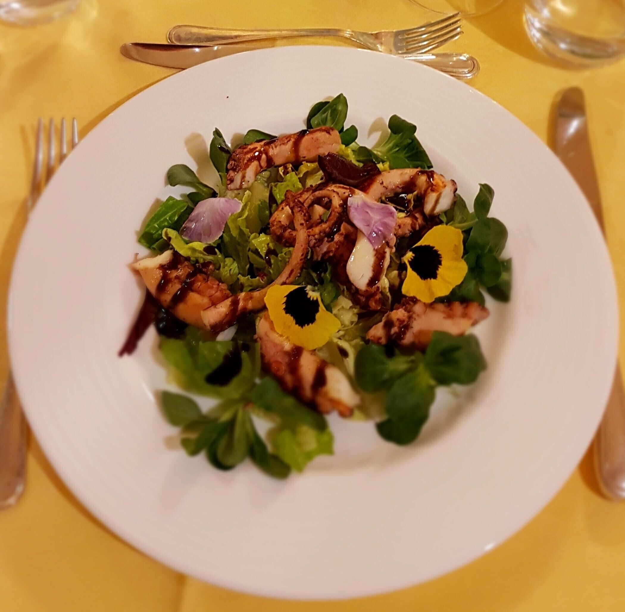 A plate of grilled octopus salad with greens and flowers on a white plate.
