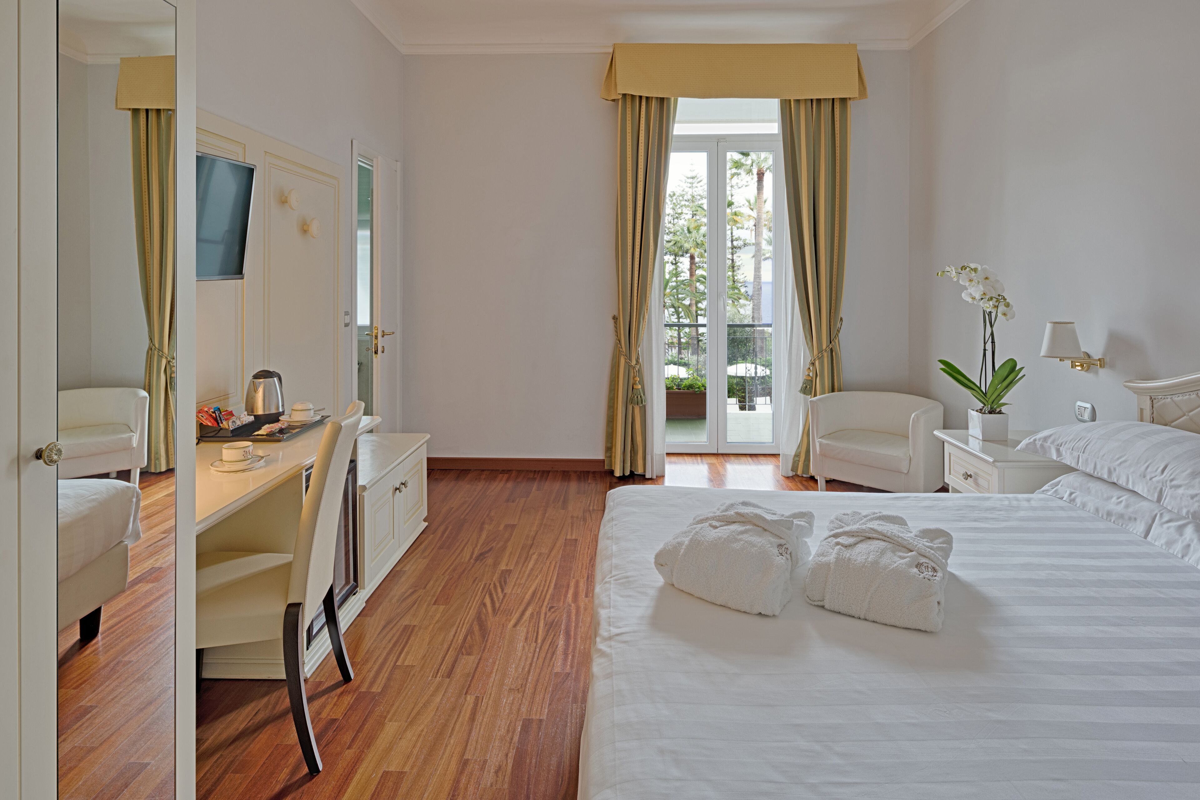 Habitación de hotel con cama, escritorio, silla, televisión y balcón con cortinas.